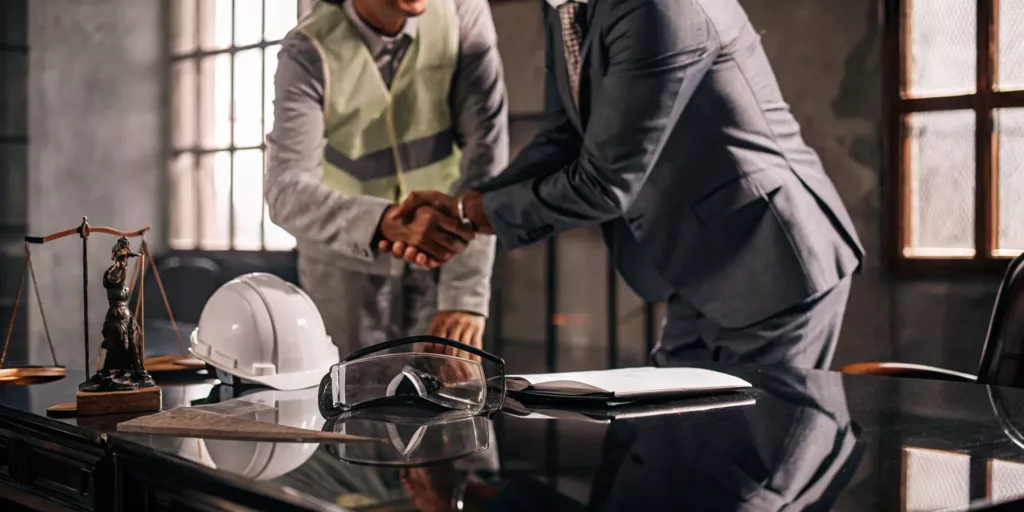 Best workers compensation lawyer and client shake hands with a safety helmet and legal scales on the desk.